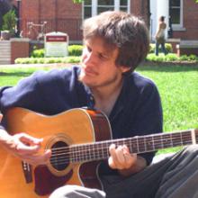 photo of Jon Watts sitting on the grass on a sunny day, playing guitar and singing