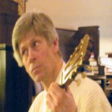 photo of Tom Allen playing guitar in front of what appears to be a pile of wooden crates.