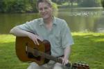 photo of Ben Tousley sitting by a lake and holding his guitar on his lap.